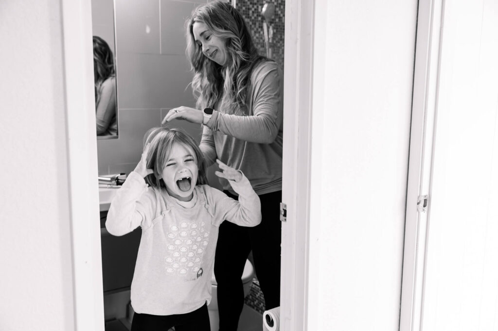 A little girl in the bathroom smiling and running away as mom tries to do her hair