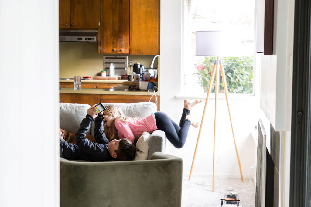 Boy laying on couch looking at his ipad, little girl hanging over arm of couch above brother, licking his arm in family documentary photo