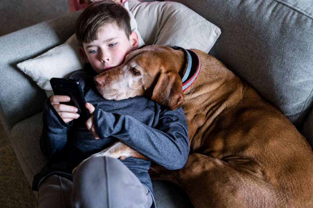 Boy laying on the couch looking at his phone with his dog's head snuggled over his shoulder and leg hugging him around the belly
