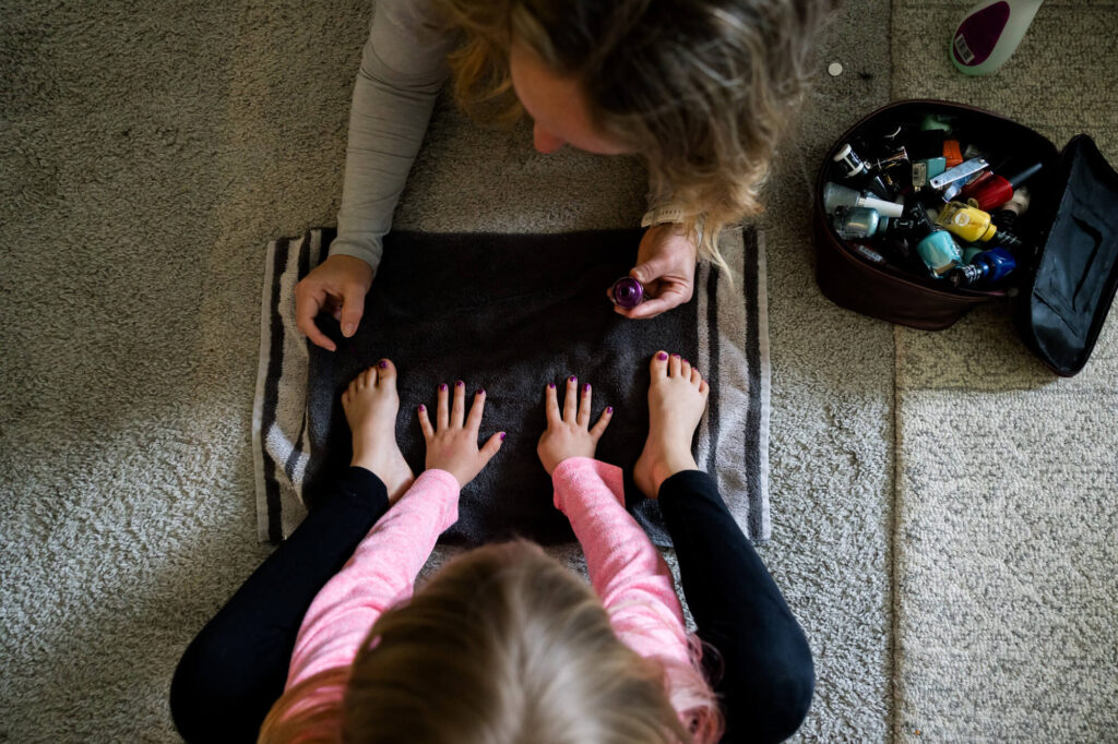 A documentary style portrait looking from above at a mom and daughter sitting on the carpet and the mom painting her finger and toe nails