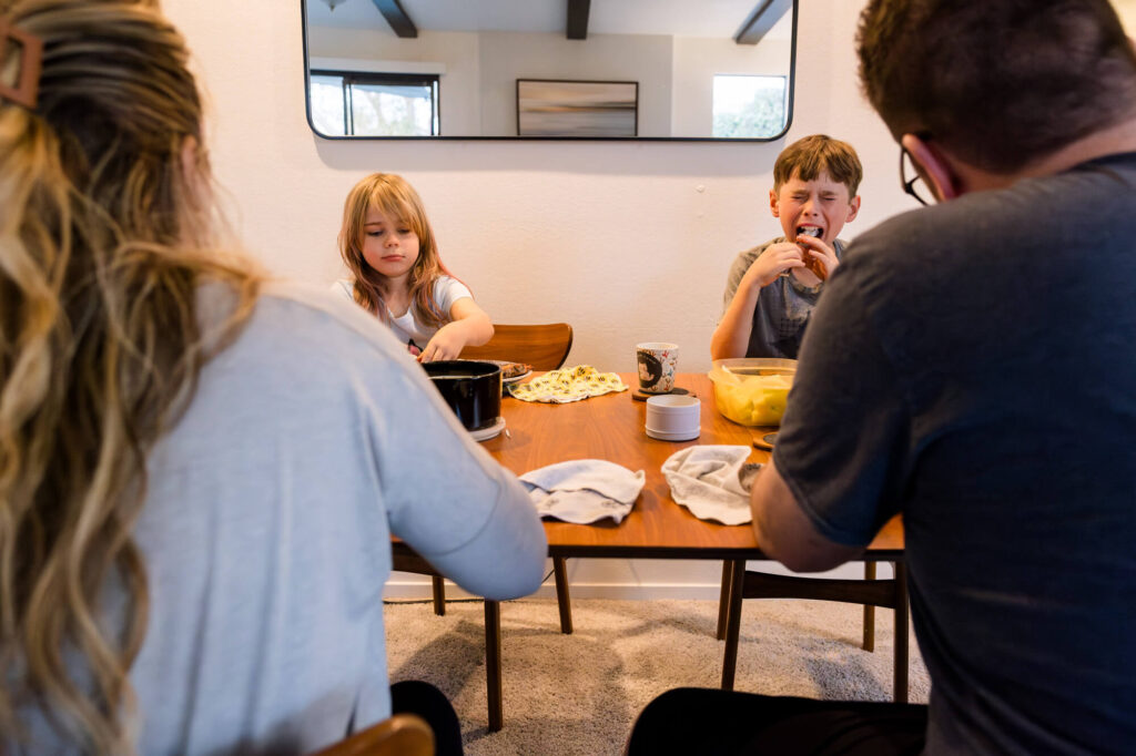 Two kids eating dinner, the son with a mouth open wide for a bite, scaffolded on either side by the backs of their parents