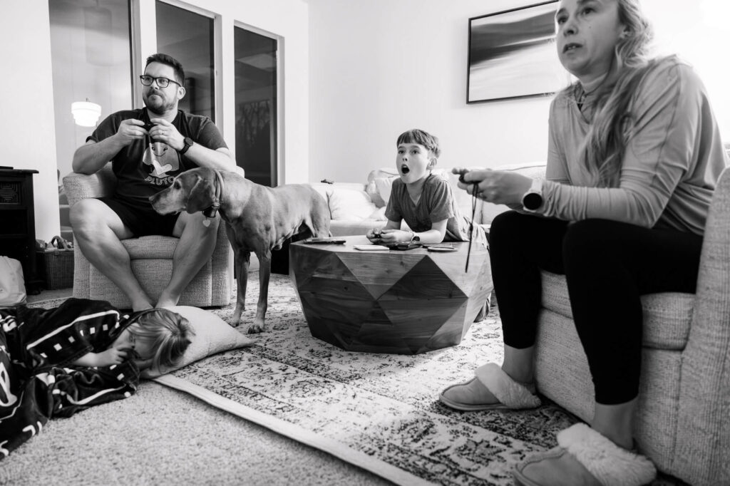 A dad, mom and son playing a video game together, the son with his mouth wide in surprise and the daughter laying asleep on the floor and the dog standing with his head in the dad's lap