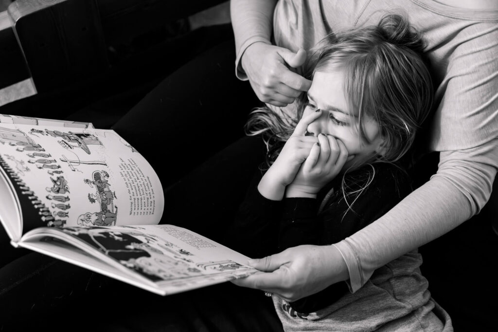 Daughter lying in mom's lap, sucking thumb, looking at a book mom is holding while mom strokes daughters hair